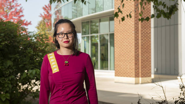 A woman in a red shirt and glasses standing in front of a building and some bushes.