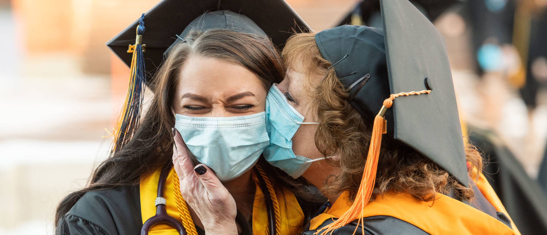 Women in masks and graduation caps exchange a kiss on the cheek.