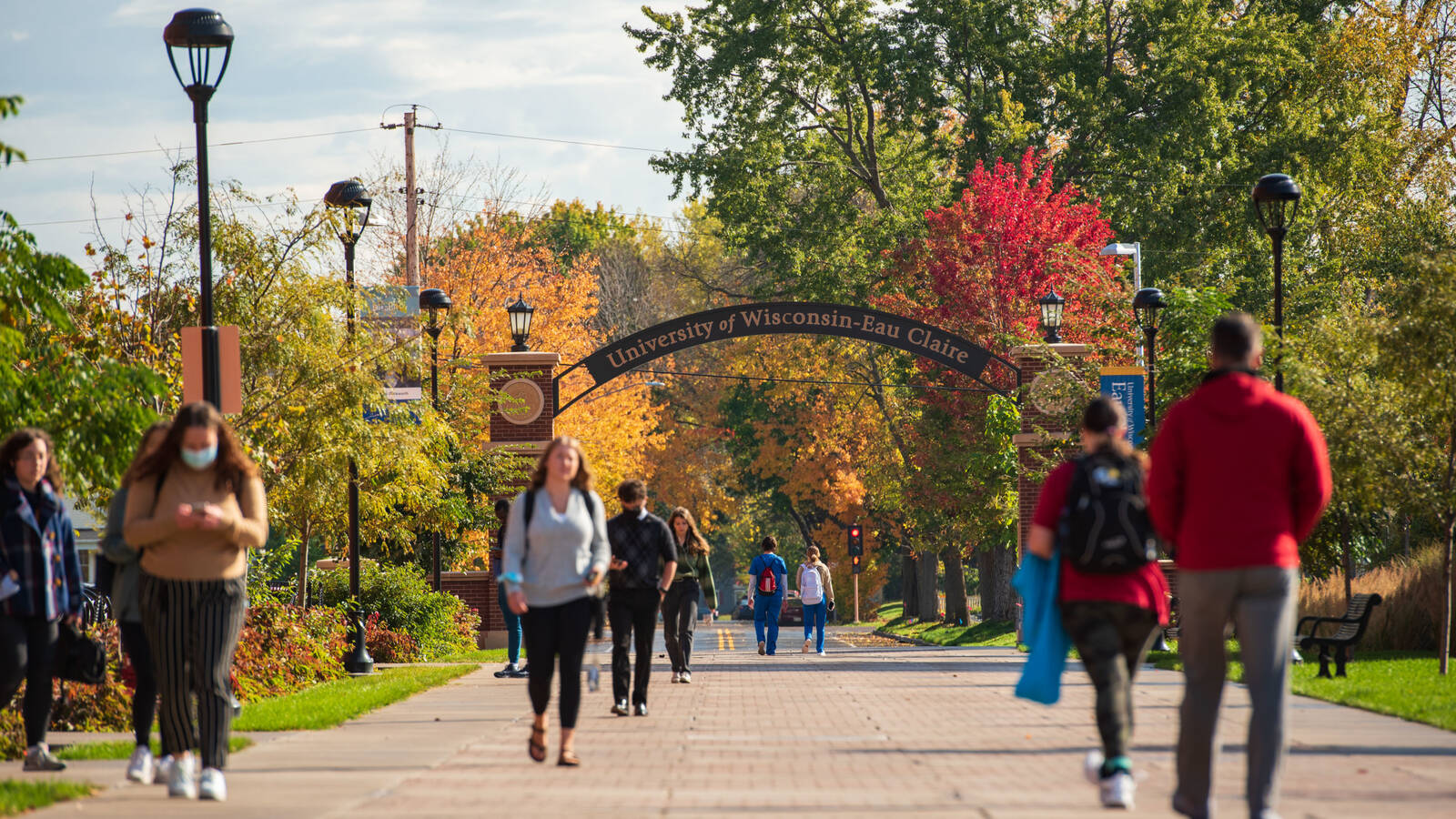 Students walking on walkway among fall leaves with University of Wisconsin - Eau Claire arch in background.