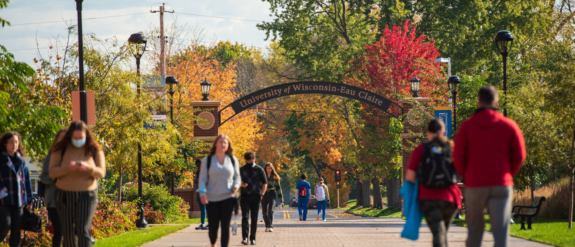 Students walking on walkway among fall leaves with University of Wisconsin - Eau Claire arch in background.