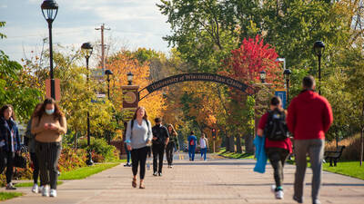 Students walking on walkway among fall leaves with University of Wisconsin - Eau Claire arch in background.