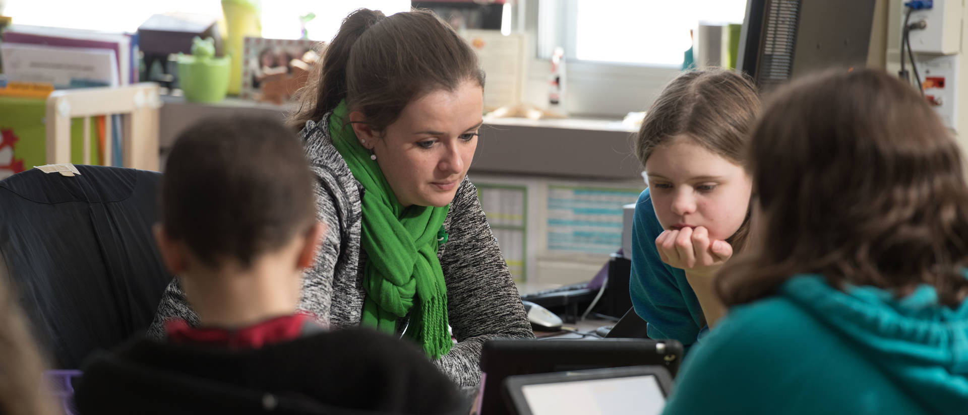 Teacher sits with students in a classroom using an iPad.