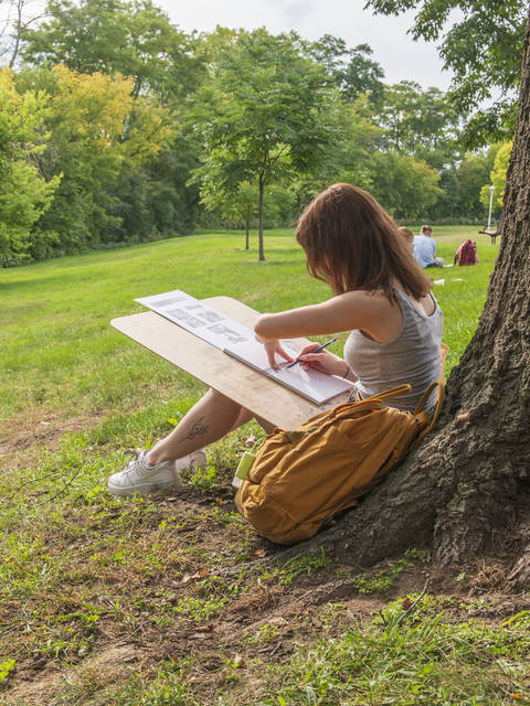 An art student draws on a clipboard next to a tree on campus.