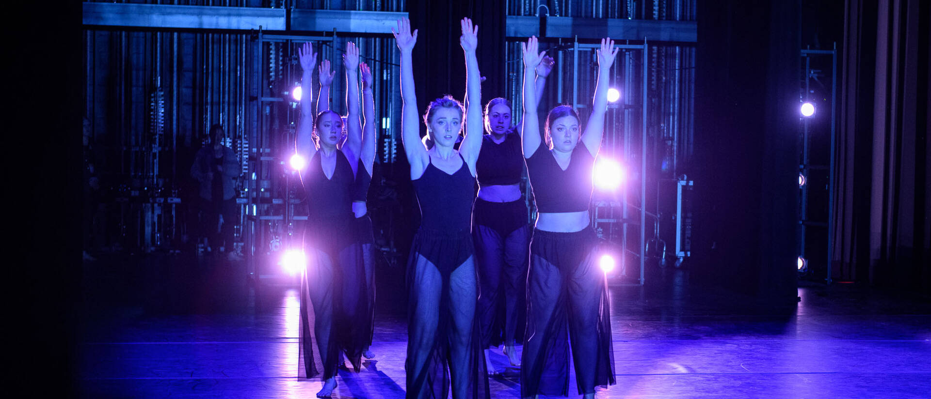 A group of students dance on stage wearing all black with spotlights behind them