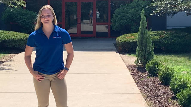 female student standing in front of commercial building, blue shirt says Rust-Oleum