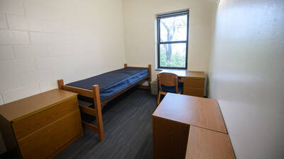 Single bedroom in Chancellors Hall apartment with bed, window, desk, chair, bookshelves, and dresser.