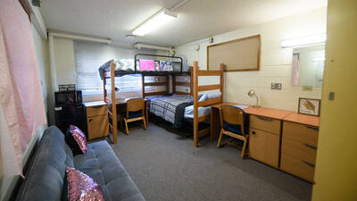 A room in Governors Hall with two bunked beds, two desks, a mini fridge and a couch.