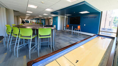 Game room in Towers Hall with colorful chairs, shuffleboard, and large TV.