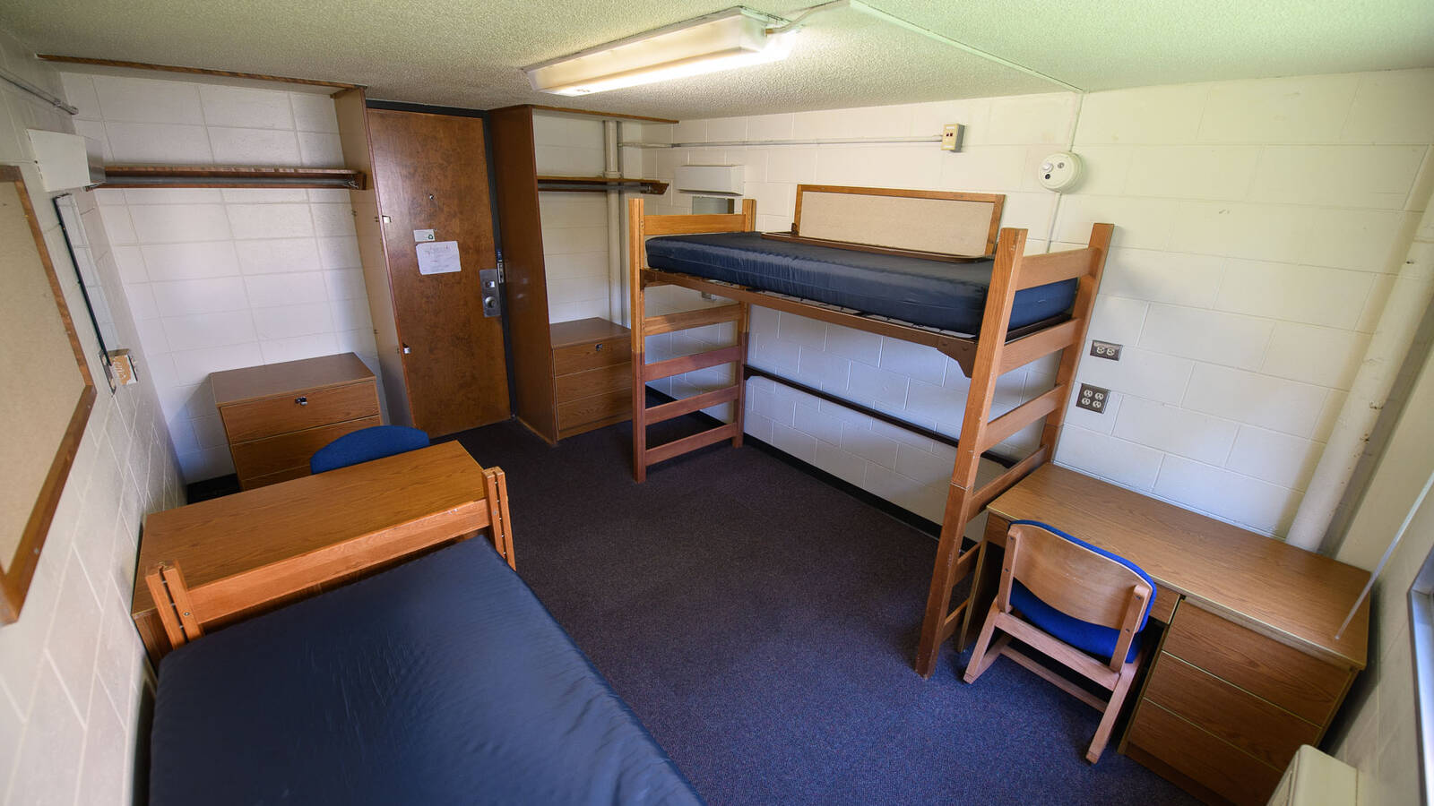 Murry Hall room (viewed from the wall opposite the door) with two beds, chairs, desks, dressers, and closet hanging space.