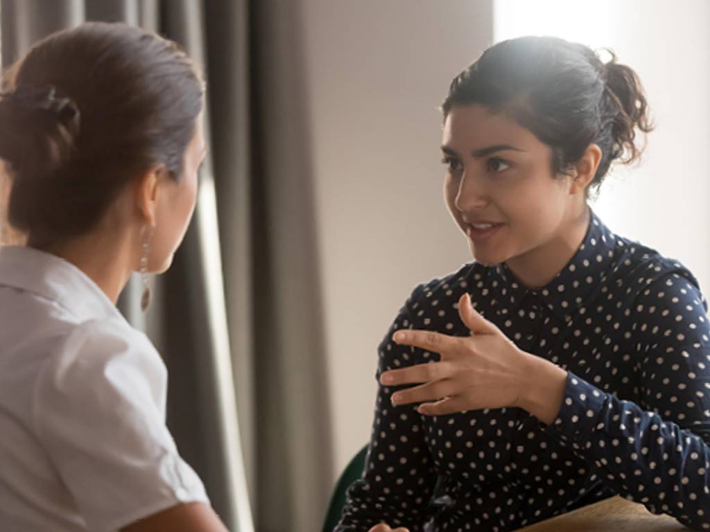 Two feminine presenting individuals are seated face-to-face; they are having a serious conversation. One is wearing a blue polka dot blouse, is facing the camera, and gesturing to the other, who's wearing a white shirt with their back to the camera.