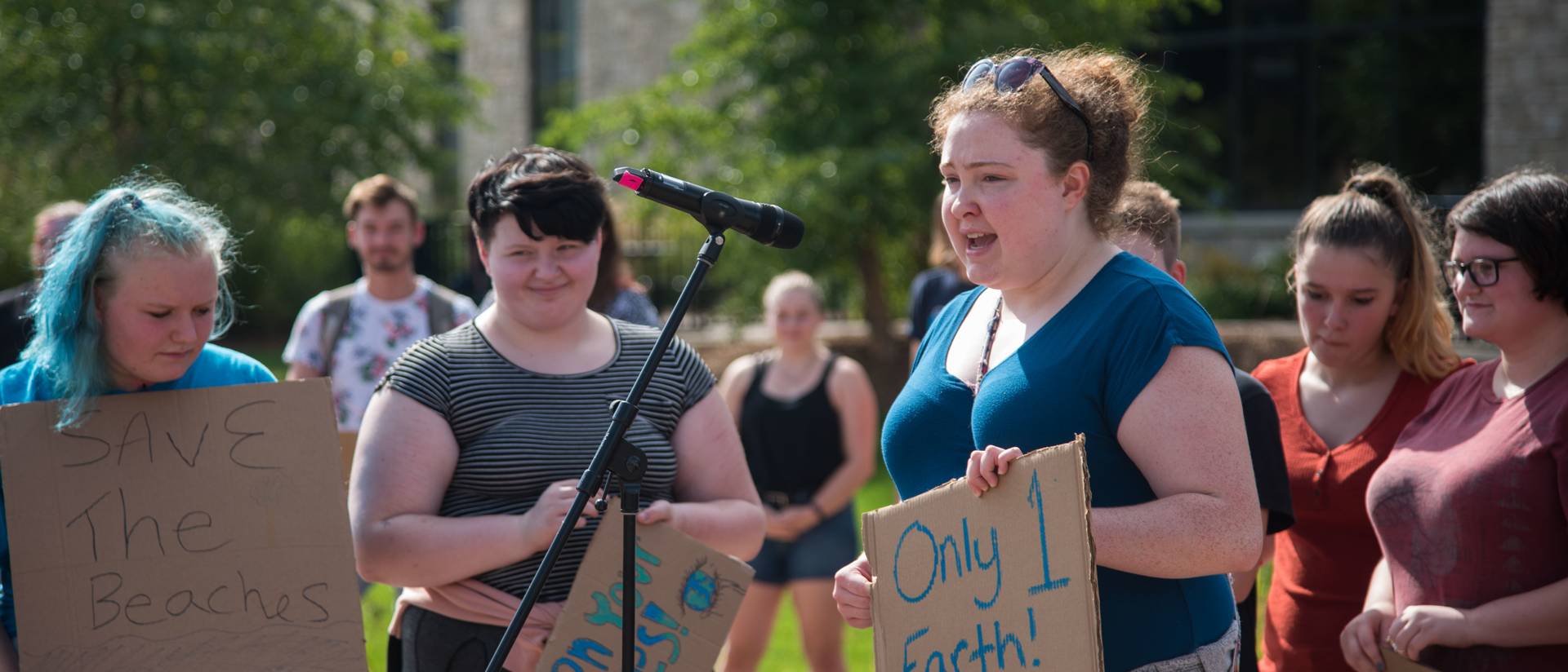 A Student holds a cardboard sign which reads "only 1 earth! Keep it safe!" while speaking into a microphone.
