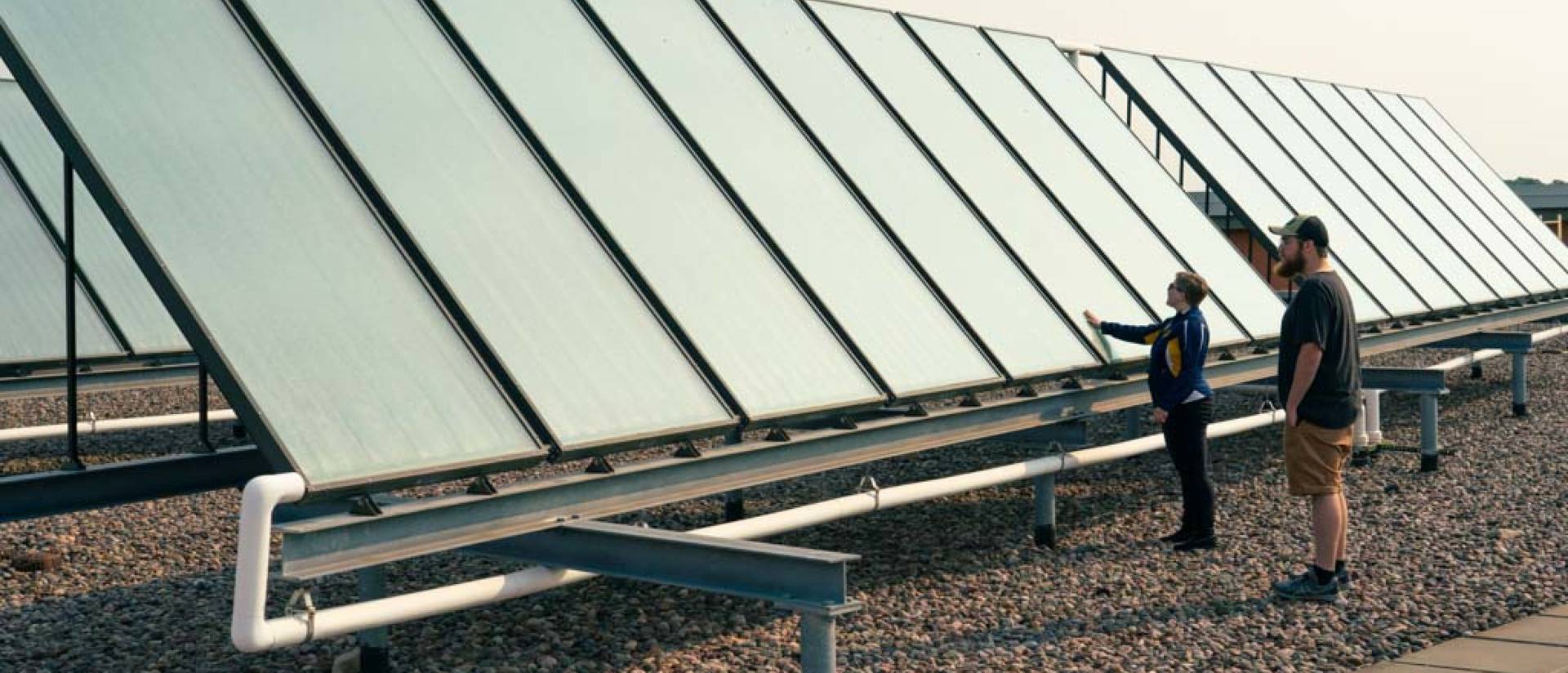 Staff stand next to the solar water heater on the roof of Davies Center. The heater is two long black rectangular panels reaching across the roof.