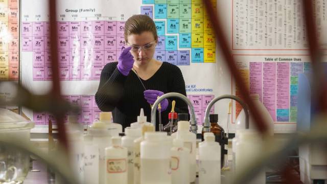 A student works with lab instruments with the periodic table hanging on the wall behind.