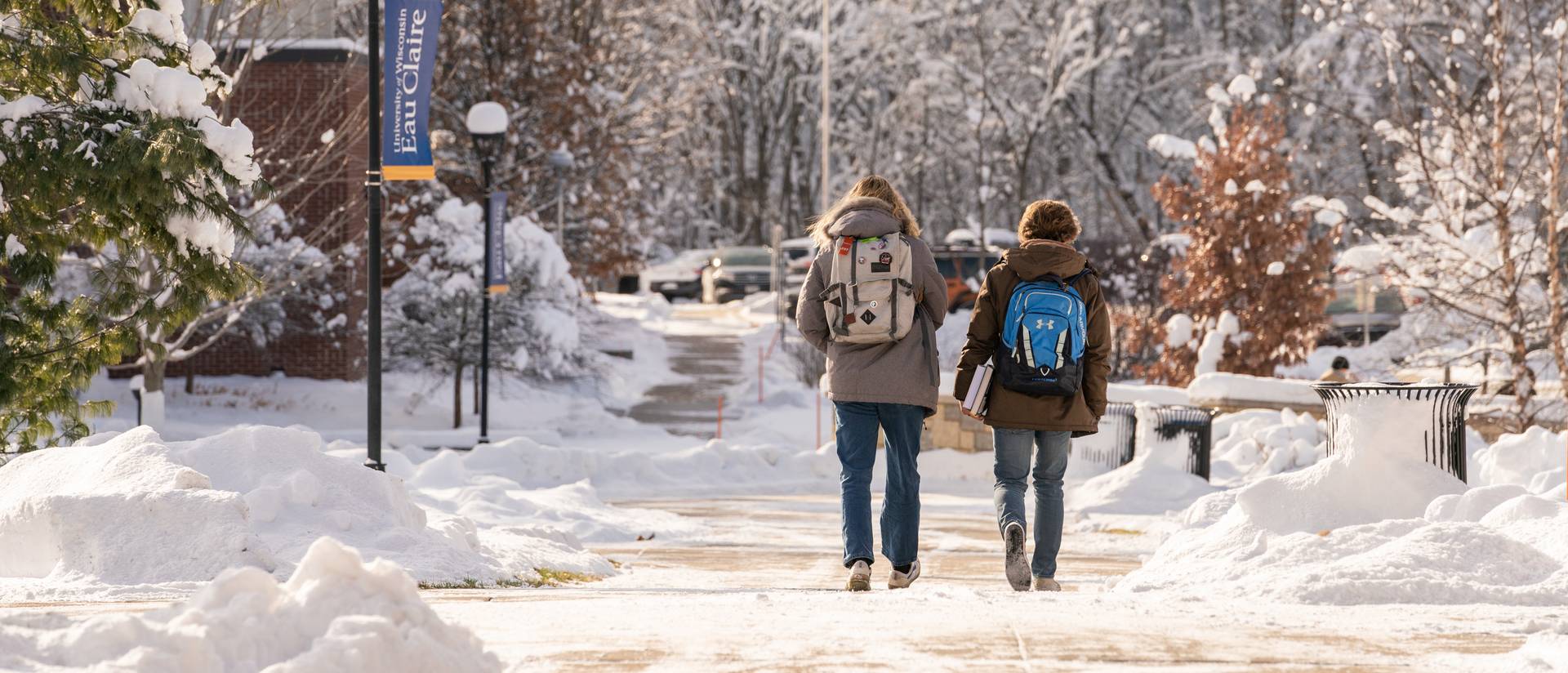 Two students walk through snow-covered campus with backpacks in tow.
