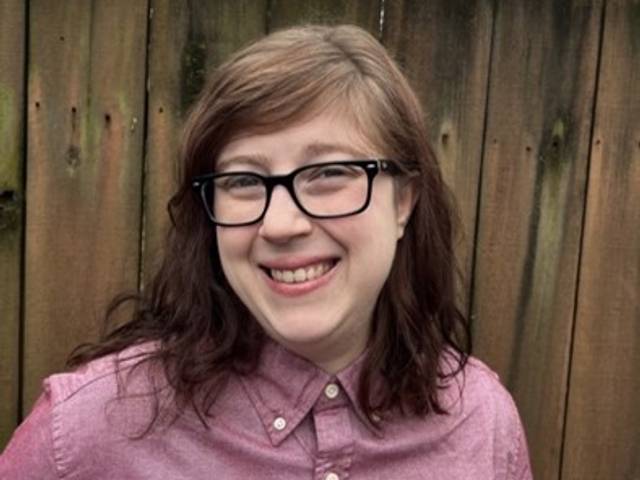 A woman in glasses smiling in front of a wooden fence.