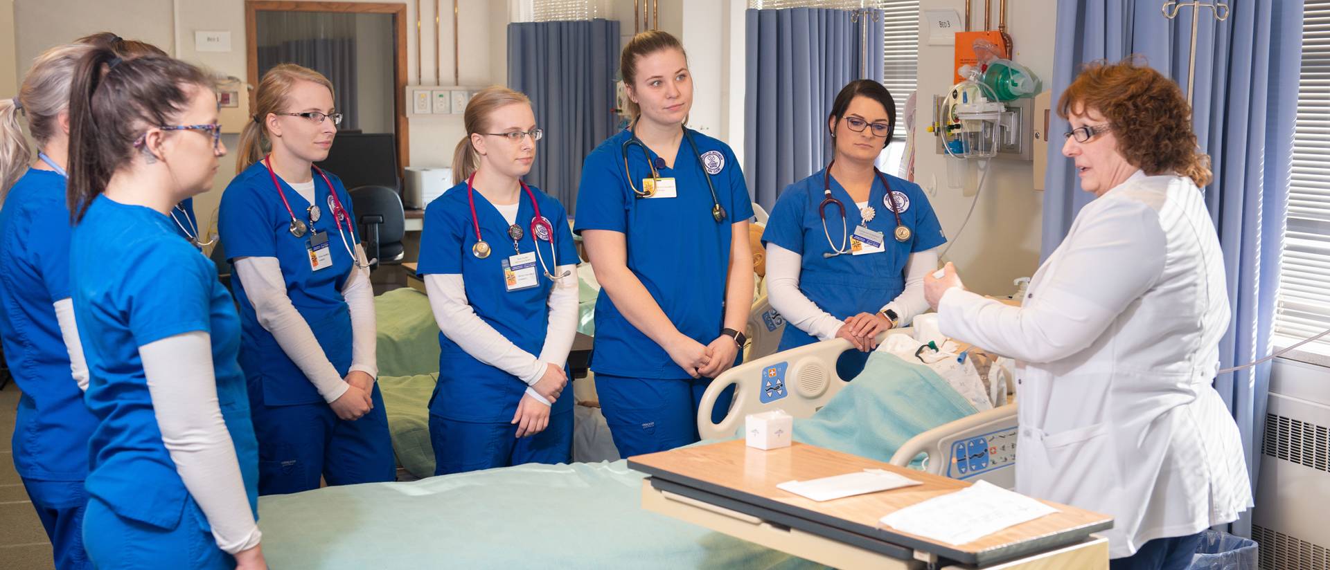 Nursing students standing around a hospital bed looking at the instructor demonstrating with a patient.