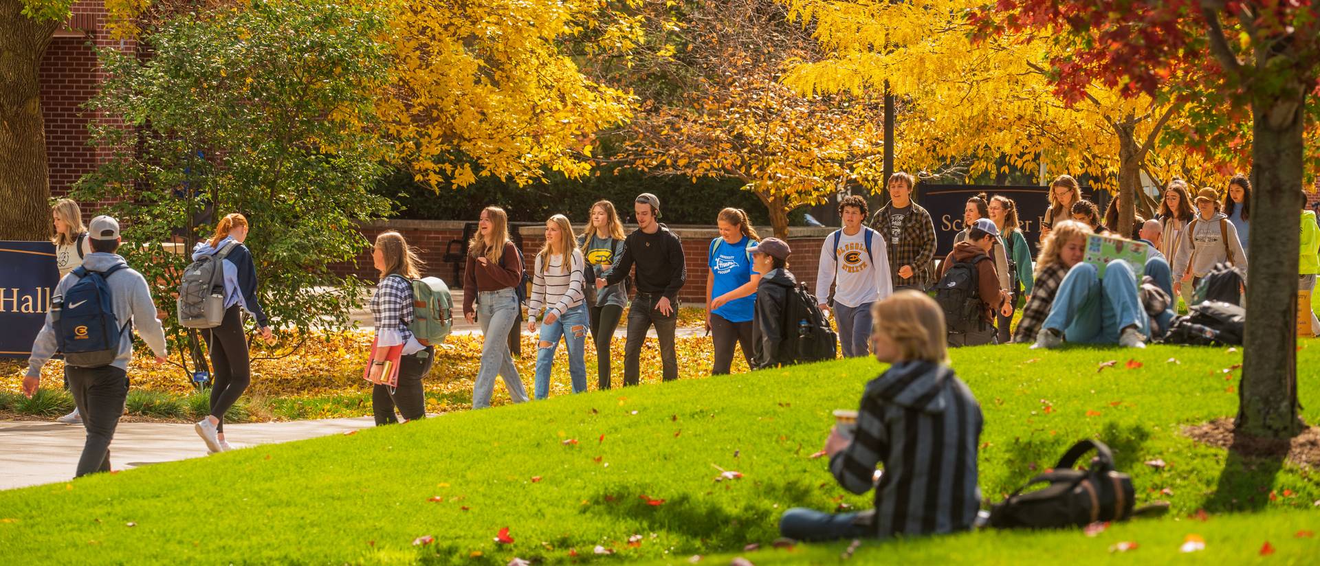 Students walk on a campus sidewalk among changing fall leaves on a sunny day