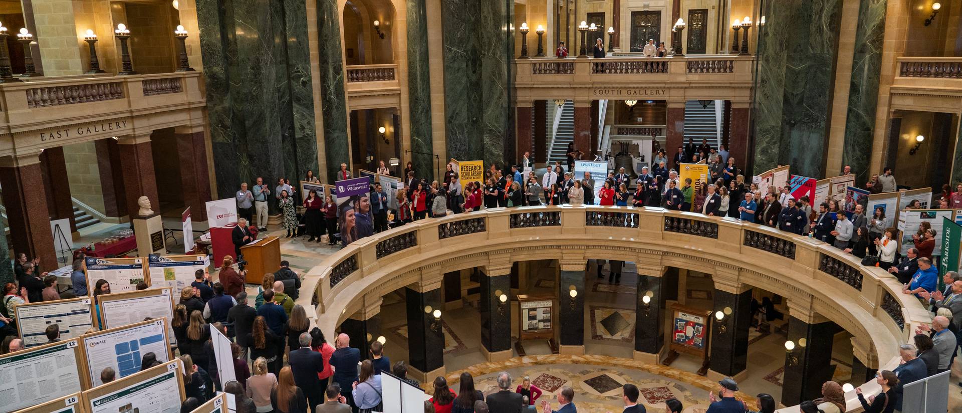 Overhead view of Madison capitol rotunda full of people and student poster presentations, speaker at a podium on left side of rotunda