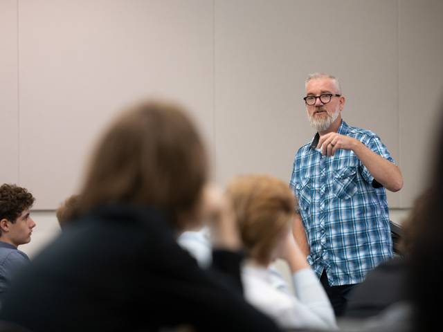 Male instructor in front of a class, blue plaid shirt