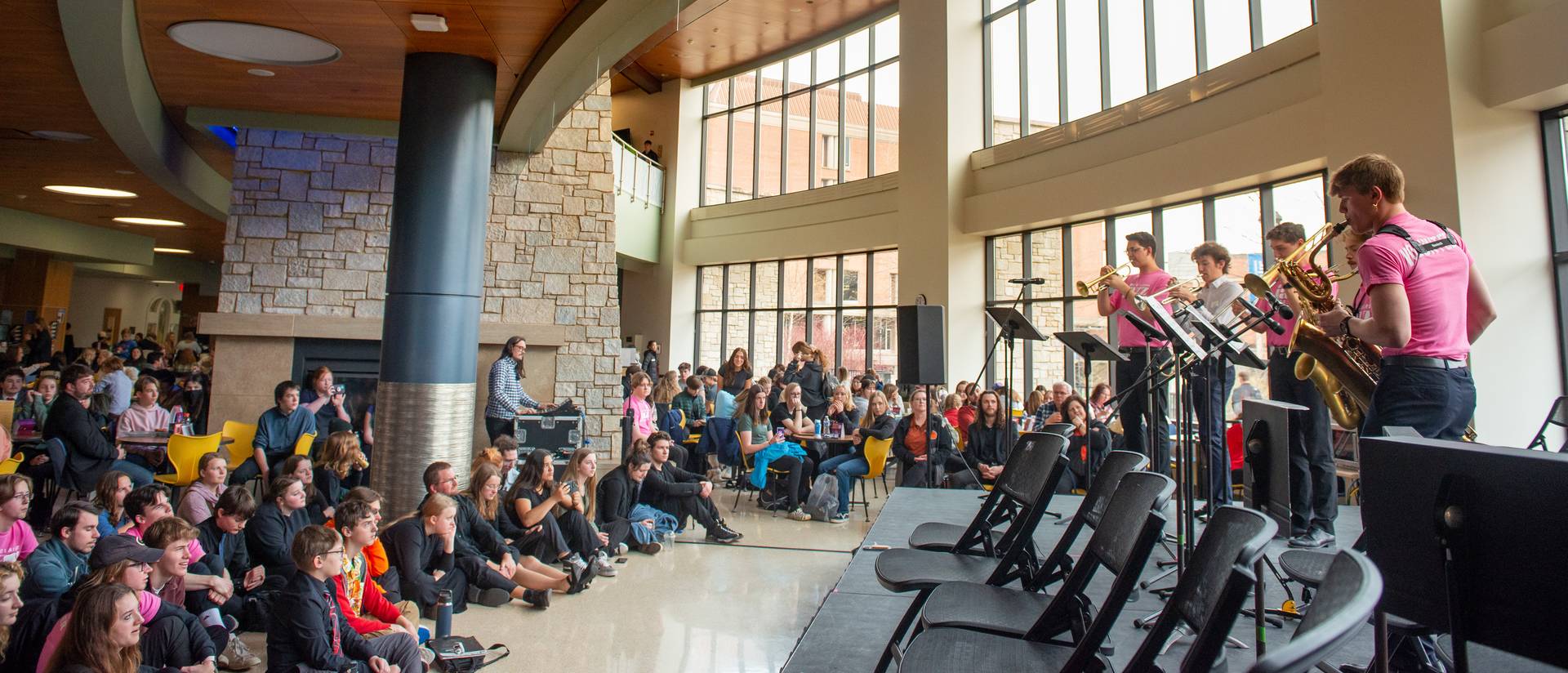 Horn players performing to a crowd in the student center, tall glass window wall behind them.