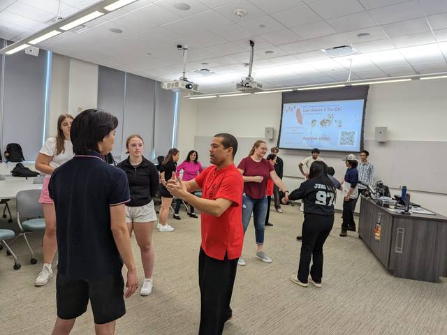 Students standing around a room listening to a Tai Chi instructor