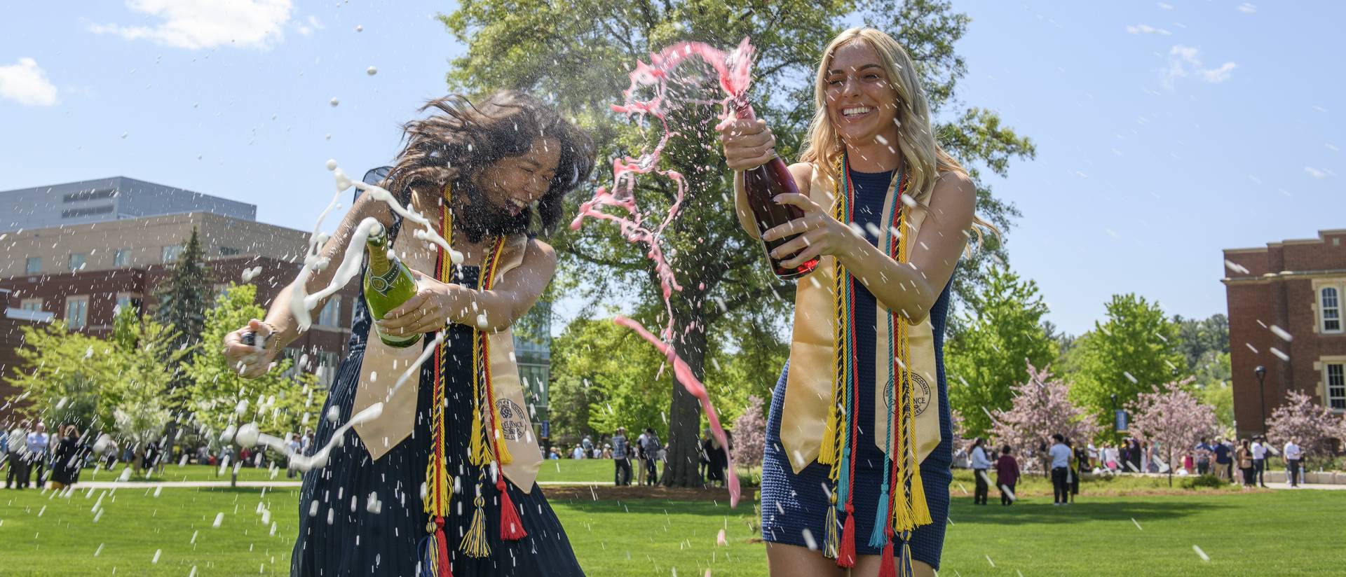 two women spraying bubbly outside on a sunny day, graduation gowns.