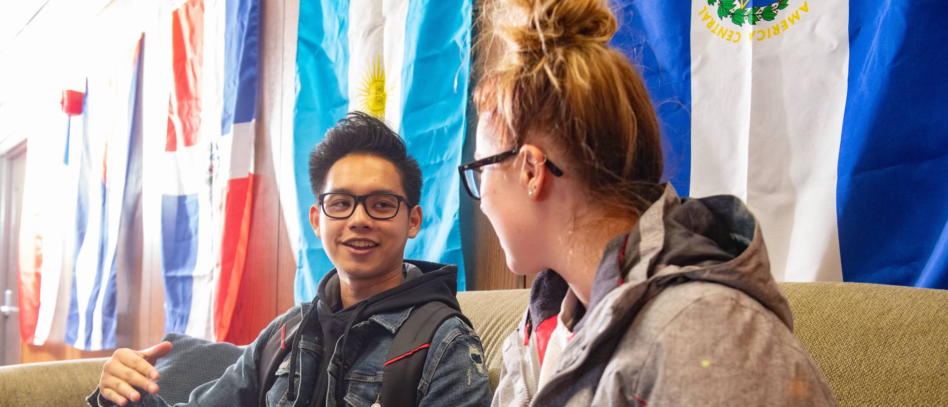 Two students chat in front of flags.