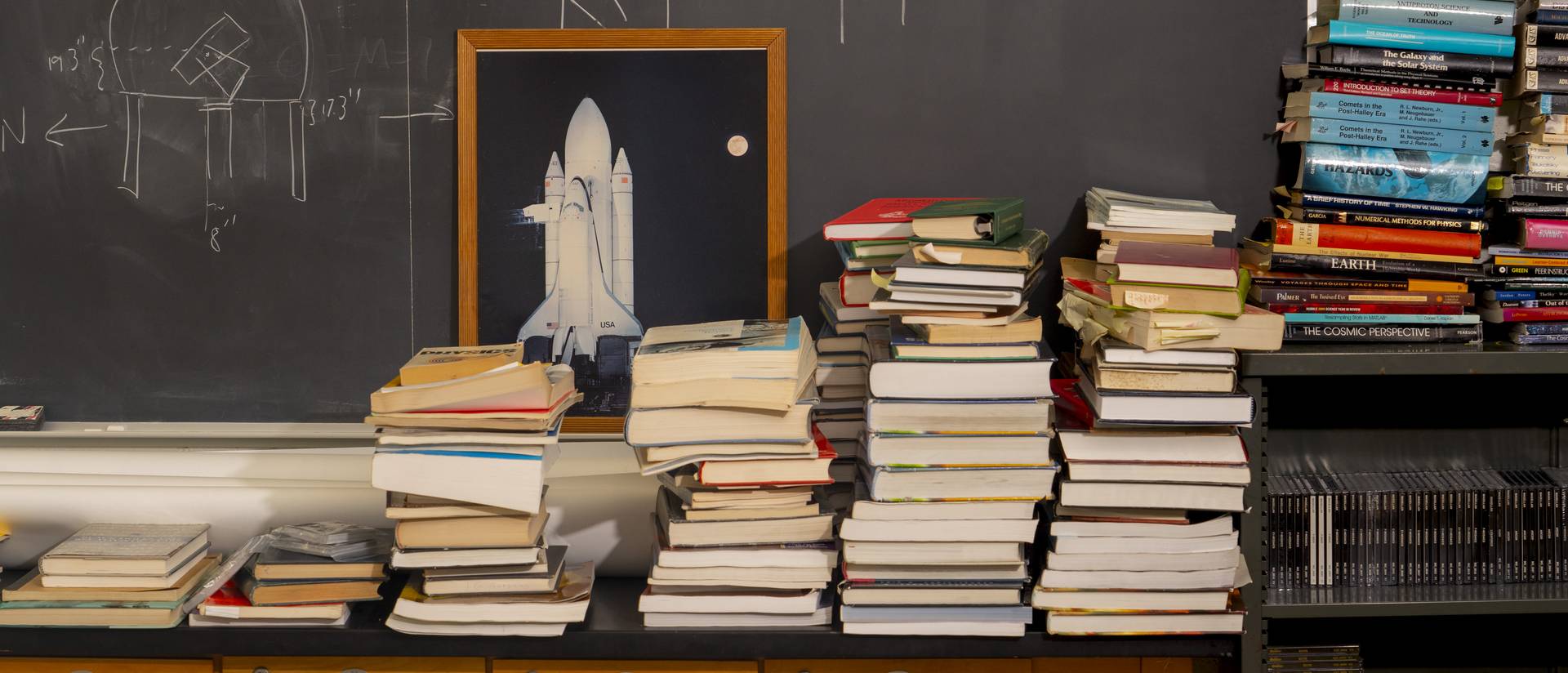 A stack of physics textbooks in front of a chalkboard and an image of a rocketship.