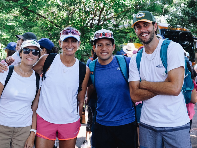 three adults posing for a group photo outdoors, man on right wearing a Packer cap