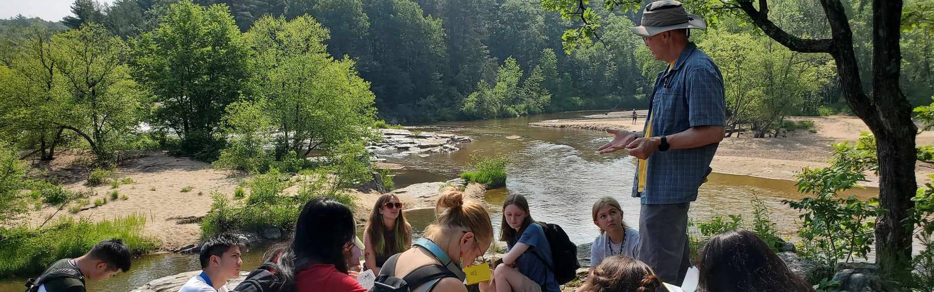 An instructor stands and talks to students as they sit on rocks near aa body of water