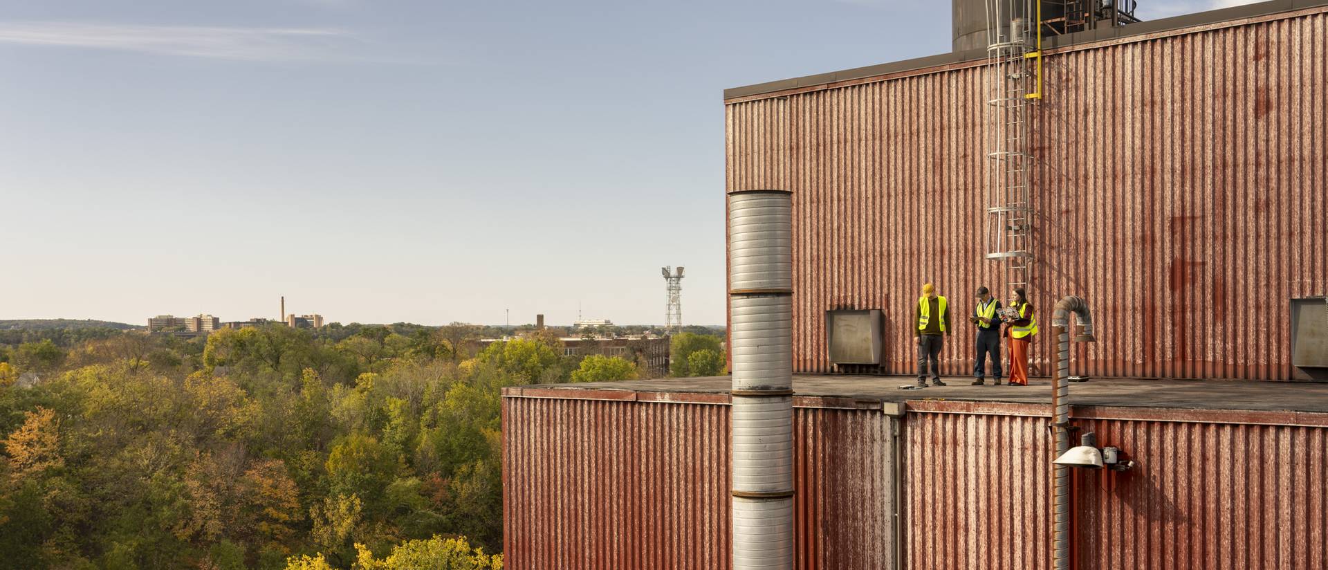 Three people wearing high-visibility vests stand on a large building's roof and collect noise measurement research data