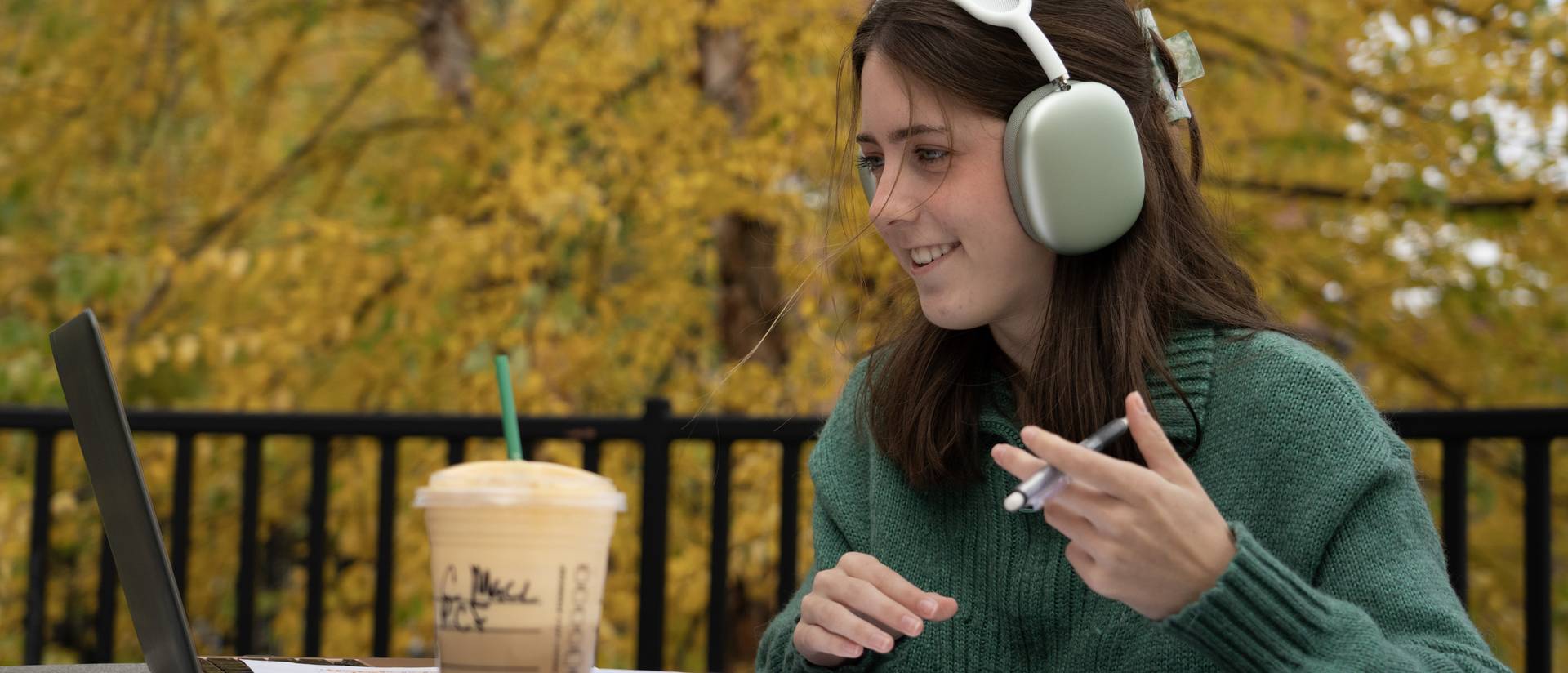 female student on the campus patio at a tble using laptop and headphones, smiling