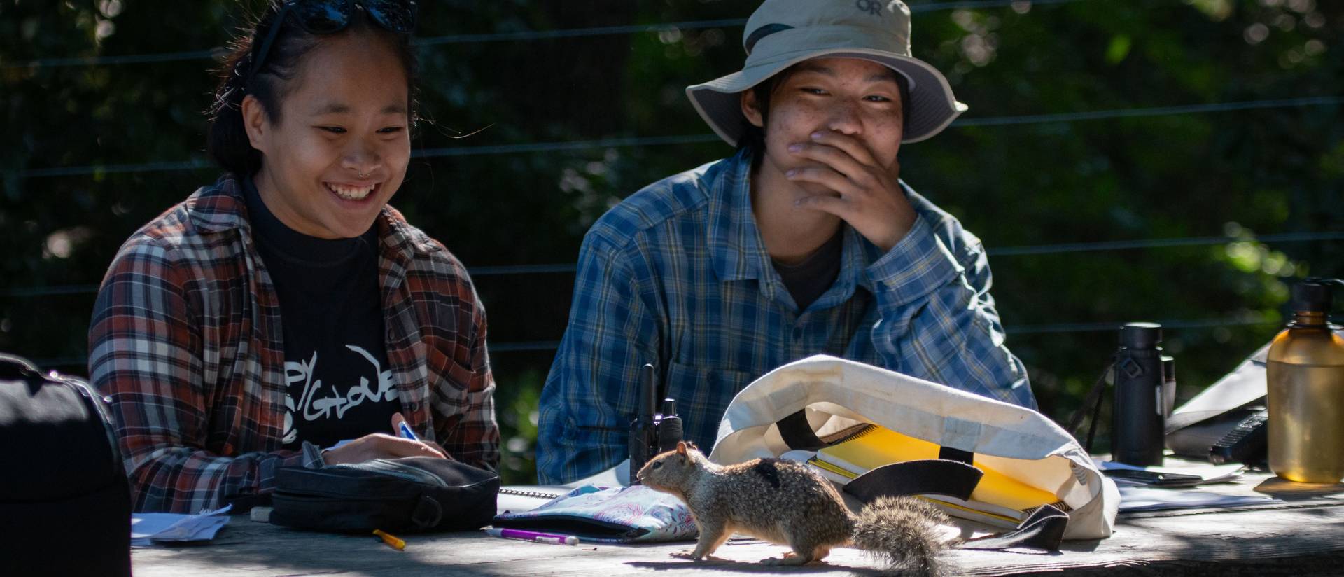 two students working at an outdoor table, a squirrel is on the table; surprised and laughing faces on the students