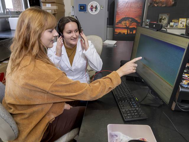 Two female students sitting side by side, in lab coats, looking at a monitor screen, one is pointing at a spot on the screen