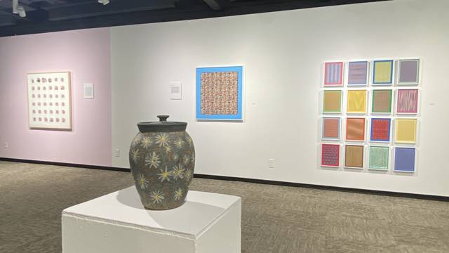 Installation view of foster gallery. A lidded ceramic pot with daisies sits atop a pedestal with a grid of framed artwork hanging on the wall behind it.