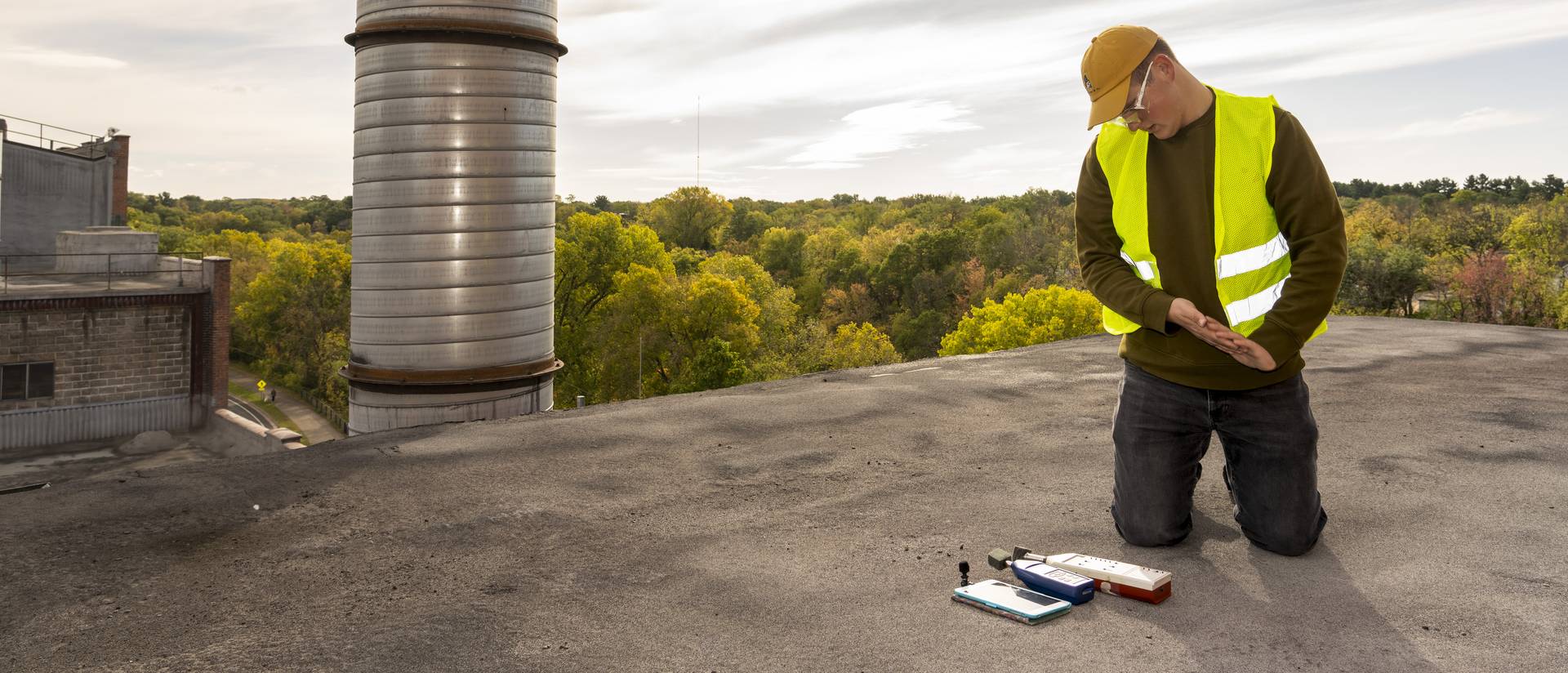 A male student wearing safety gear kneeling on a metal rooftop checking some measurement instruments