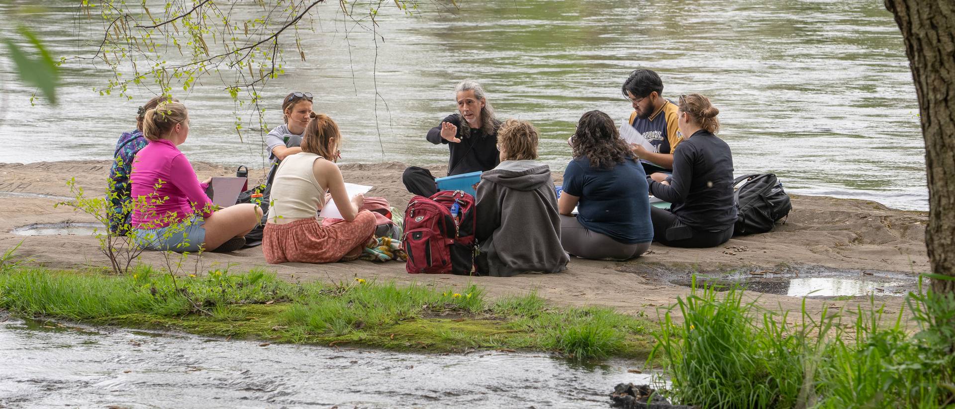 Students sit in a circle and listen to their professor speak while they sit on a rock near the river.
