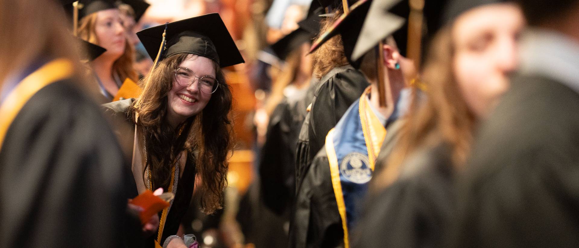 A graduate smiles at the camera between rows of graduates in caps and gowns.