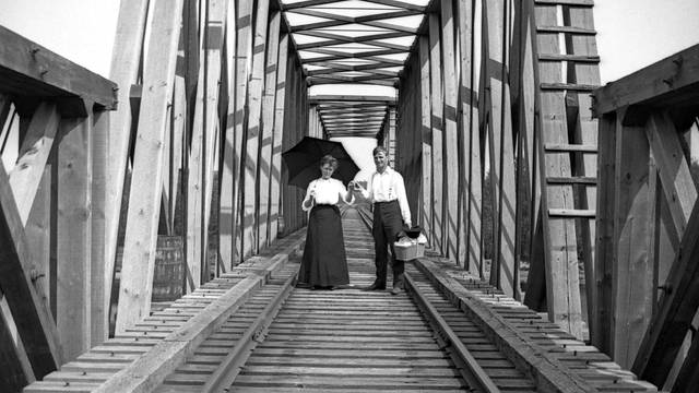 Two people stand on a wooden bridge, with one holding an umbrella, and a single track below them.