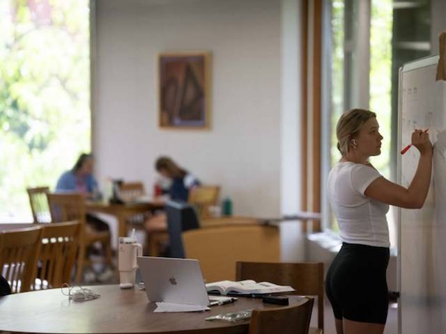 Woman writing on whiteboard as others study in the background