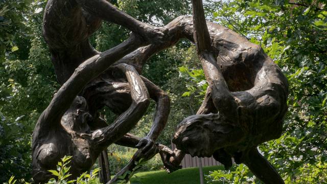 The image depicts an outdoor sculpture of three intertwined human figures that appear to form a sphere. The sculpture is bronze, giving it a polished and slightly reflective surface.