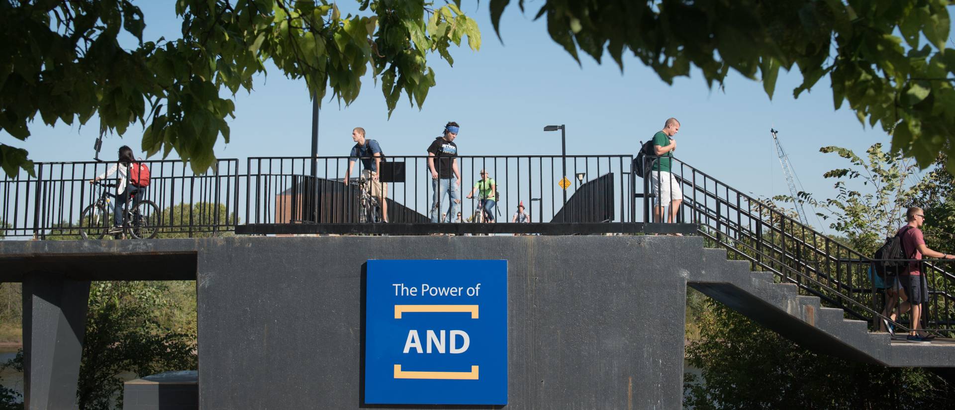 Students walk and bike on the campus bridge, with "The Power of AND" logo displayed at the end of the bridge.