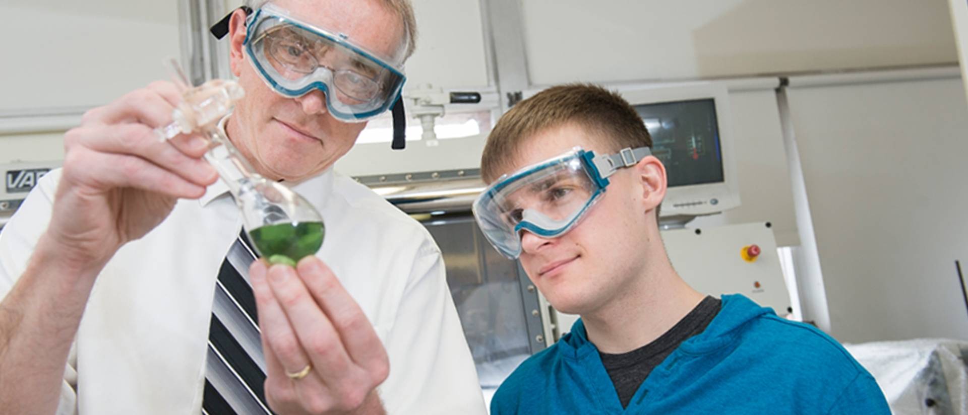 A professor and student wear safety goggles as they observe a green liquid held in a glass.