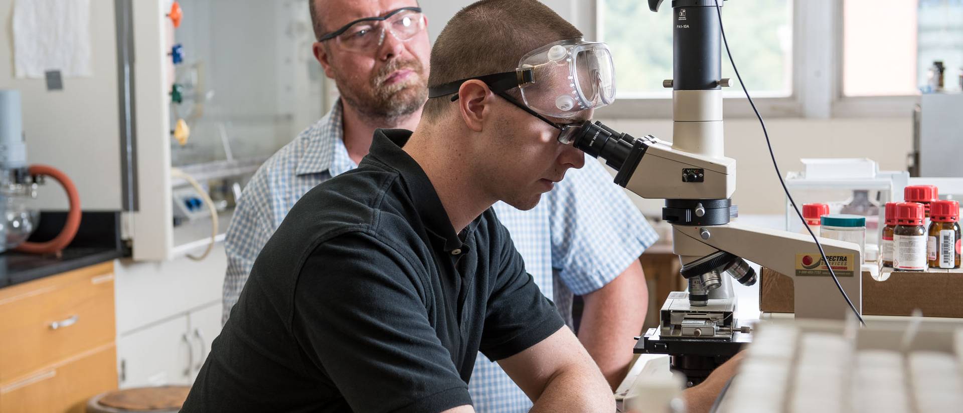 An instructor watches a student as they use a microscope.