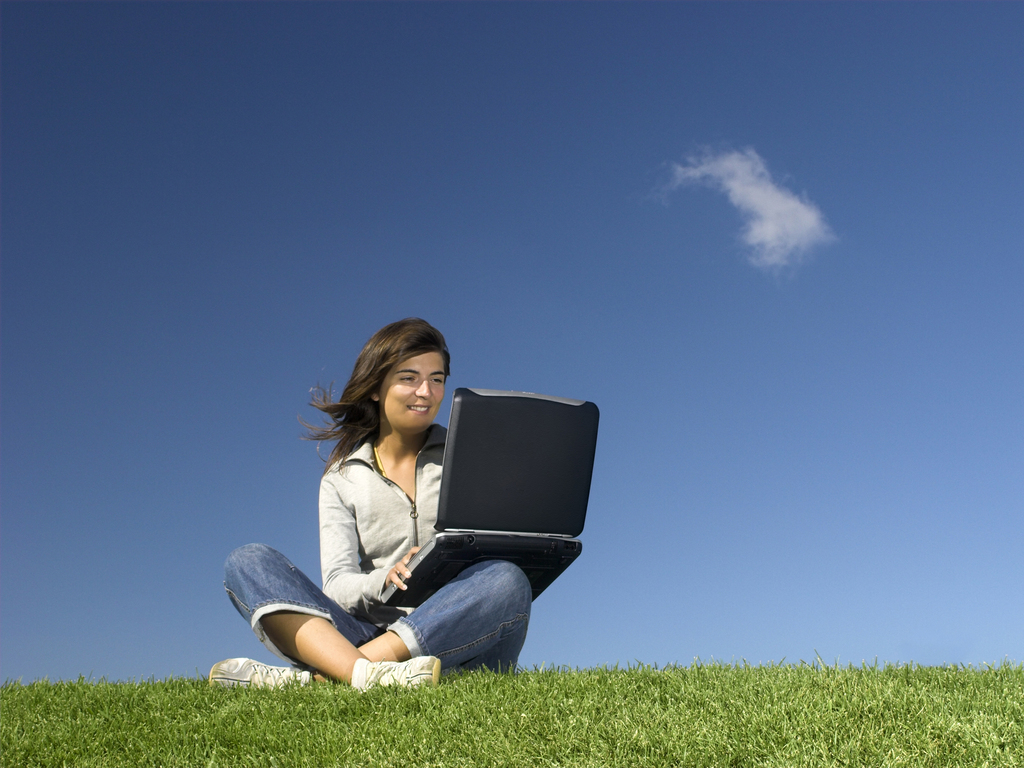Adult woman sitting cross legged in grass with laptop set to a blue ski background. 