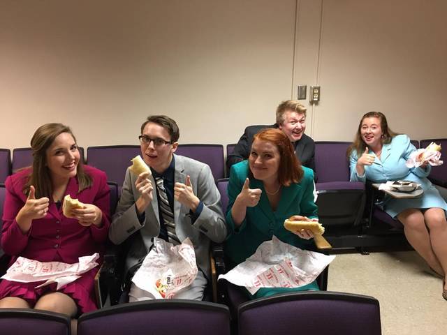 Five professionally dressed students smile and give a thumbs-up as they take a lunch break.