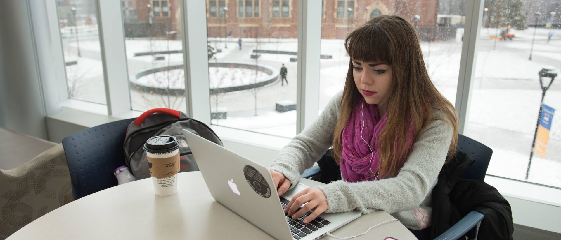 A student sits near a window typing on their laptop in Centennial Hall with snow falling outside.