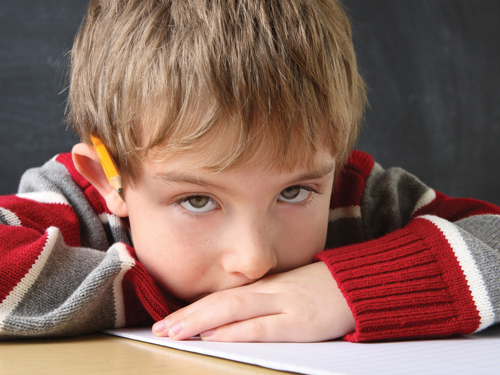 A young boy lays with his head on his arms looking at the camera, pencil tucked behind his ear.