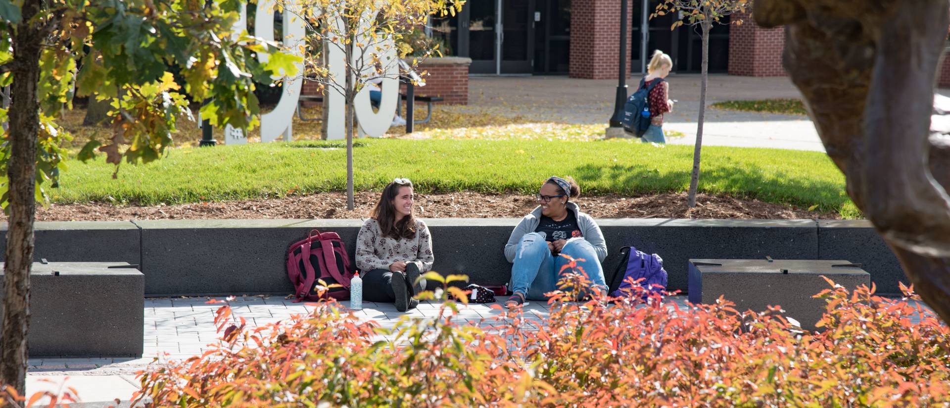 Two students sit and laugh together on the Campus Mall in the Fall.