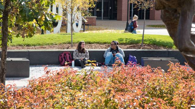 Two students sit and laugh together on the Campus Mall in the Fall.